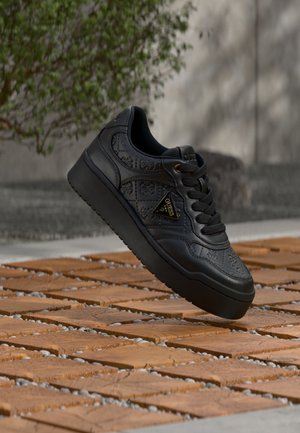 Sneaker platform nera con motivi floreali in rilievo e logo triangolare dorato Guess sul lato, posizionata sopra un pavimento in legno.