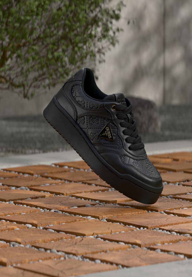 Μαύρο sneaker πλατφόρμα με ανάγλυφα λουλουδένια σχέδια και χρυσλό τριγωνικό λογότυπο Guess στο πλάι, τοποθετημένο πάνω από ξύλινο πάτωμα.