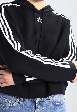 Person wearing black Adidas hoodie with white stripes on sleeves and logo on chest, arms crossed, paired with blue jeans.