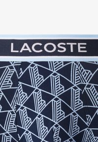 Bóxer azul marino con un patrón geométrico en azul claro. Cintura elástica con el logo "LACOSTE" en blanco. Textura de tela suave.