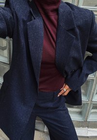 Oseba v mornarsko modrem suknji s širokimi reverji, oblečena v burgundski pulover s turtle vratom, roka položena na boke, kjer se vidi srebrn prstan.