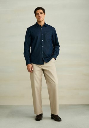 Man wearing dark blue buttoned shirt, beige trousers, and dark brown shoes standing against a plain beige wall.