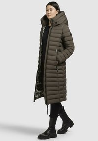 Long olive green puffer coat with a hood, featuring horizontal quilting, front zipper, and side pockets. Worn over black leggings and boots.