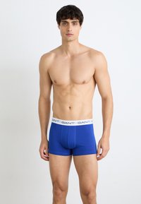 Boxeri albaștri dintr-un material elastic, cu un brâu elastic alb pe care este imprimat de mai multe ori numele mărcii "GANT".