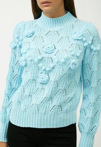 Maglione lavorato a maglia di un azzurro chiaro con collo alto, caratterizzato da decorazioni floreali e un motivo testurizzato. Maniche e orlo a coste.
