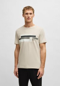 Camiseta color beige claro hecha de tela suave. Presenta elementos horizontales en blanco y negro con patrones texturizados. Detalle del logo minimalista.