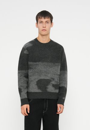Un pull gris à motifs avec un col rond, présentant un design texturé dans des nuances variées de gris et un ourlet côtelé, associé à un jean foncé.