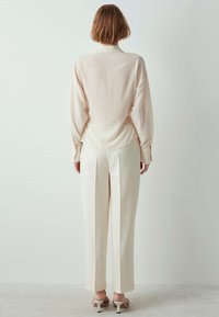Blouse texturée beige clair avec une coupe décontractée, associée à un pantalon blanc ajusté. La tenue présente un col simple et de légères rayures.