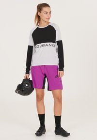 Radfahr-Outfit mit einem grau-schwarzen Langarmshirt und lila Shorts. Enthält einen schwarzen Helm und schwarze Radschuhe.