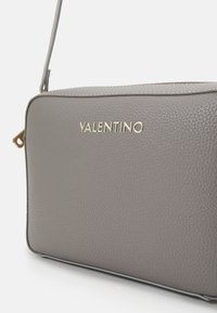Sac bandoulière en cuir gris avec finition texturée. Présente un logo doré "Valentino" et une fermeture éclair. Forme rectangulaire compacte avec sangle réglable.