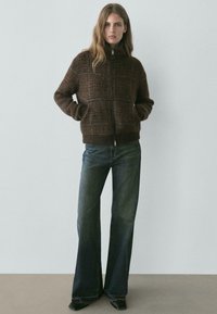 Kobieta stojąca z rękami w kieszeniach, ubrana w brązową, kraciastą kurtkę na zamek i ciemne, szerokie dżinsy na tle jednolitej ściany.