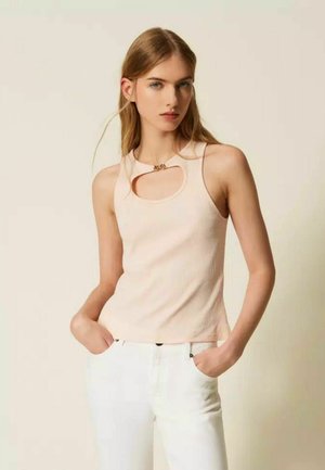 Giovane donna con capelli lunghi che indossa un top beige senza maniche con dettaglio traforato e pantaloni bianchi, in piedi con le mani nelle tasche.