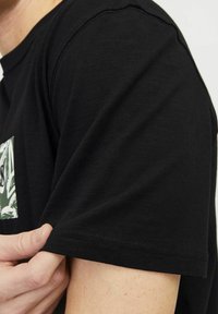 Schwarzes kurzärmliges T-Shirt aus strukturiertem Stoff mit rundem Halsausschnitt und einem grünen Grafikdesign im Bereich der Brusttasche.