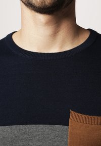 Stickad tröja med rund hals, prydd med mörkblå, grå och bruna horisontella ränder. Märkbar framficka i brunt med ribbat mönster.