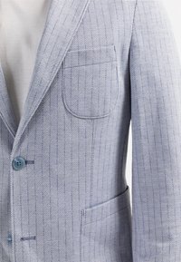 Blazer gris clair avec fines rayures diagonales bleues, deux boutons bleus visibles et une poche poitrine arrondie sur une chemise blanche.
