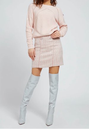 Kobieta ubrana w jasnoróżowy sweter z dzianiny, dopasowaną różową mini spódniczkę z tkaniny typu tweed z guzikami oraz białe wysokie kozaki, stojąca na tle jednolitego tła.