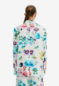 Marynarka w kwiaty z białym, teksturowanym tłem, z intensywnymi kolorami, takimi jak niebieski, fioletowy, czerwony i żółty. Tylny widok pokazuje rozcięcie.