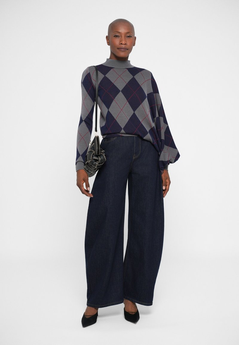 Donna che indossa un maglione argyle grigio e blu navy, jeans scuri a gamba larga, tacchi neri a punta e una borsa nera con dettagli a catena.