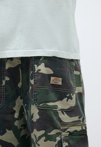 Kamuflāžas šorti tumši zaļā un bēšā krāsā ar izteiktām kabatām, auduma etiķeti aizmugures kabatā un brīvā piegriezuma dizainu.