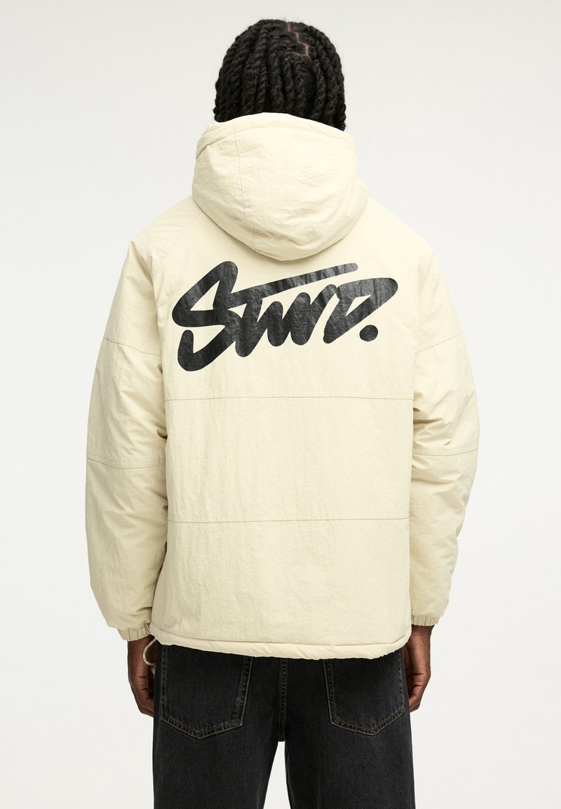 Personne aux cheveux tressés portant une veste à capuche crème avec un grand texte noir stylisé « Stmr. » dans le dos et un jean foncé.