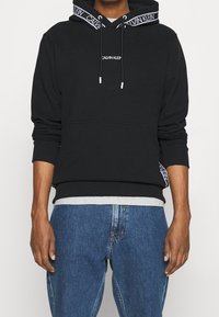 Felpa nera con logo, polsini e orlo a coste. Presenta lacci bianchi e un rivestimento a motivo nella cappuccio. Abbinata a jeans blu.