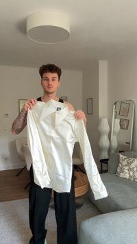 Valkoinen pitkähihaainen paita, jossa on kärkipoikkinen kaulus ja sileä pinta, pidetään ylhäällä hyvin valaistussa huoneessa, jossa on neutraalinvärisiä huonekaluja.