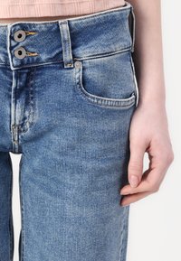 Faded blue denim jeans feature a high waist with two metallic buttons, a front pocket, and visible stitching details. Smooth texture.
