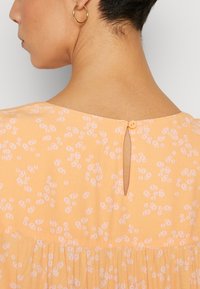 Haut en tissu pêche avec un motif floral, une ouverture en goutte d'eau à l'arrière avec un bouton, et une finition douce et texturée.