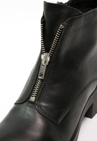 Bottine en cuir noir avec fermeture éclair argentée partiellement ouverte, montrant une doublure intérieure noire et des coutures sur une surface lisse.
