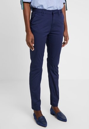 Osoba ubrana w granatowe dopasowane spodnie, jasnoniebieską koszulę oraz pasujące granatowe teksturowane mokasyny, stojąca na jednolitym tle.