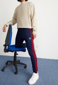 Beige sweatshirt, marinblå byxor med röda ränder, logodetalj, vita sneakers och en blå kontorsstol på ett blått heltäckningsmattgolv.
