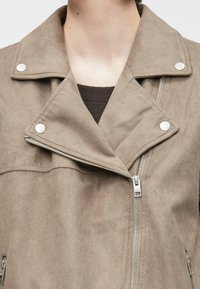 Beige faux-suede jakke med notched krage, sølvsnapknapper og skrå glidelås, som viser en jevn, teksturert overflate.