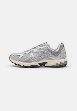 Zapatilla deportiva gris con parte superior de malla y sintético, superficie texturizada, media suela acolchada de color crema-blanco y suela negra resistente.