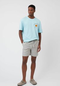 Camiseta de algodón azul claro con un gráfico del sol en el pecho, combinada con pantalones cortos a rayas grises y blancos. Calzado casual tipo slip-on completa el look.