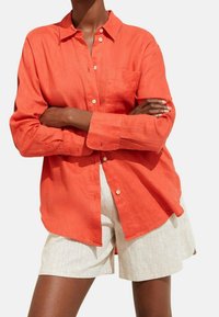 Oranges Leinenhemd mit einem Button-Down-Kragen, das eine Brusttasche und lange Ärmel aufweist, kombiniert mit beigen Leinen-Shorts.