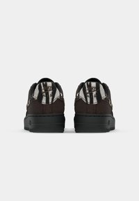 Zapatillas marrones con acentos negros, con una parte superior de tela texturizada y una suela gruesa negra. La parte trasera muestra un diseño gráfico en blanco y negro.