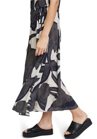 Femme portant une robe maxi à motifs noirs et blancs, tenant la jupe, marchant en sandales plateformes noires sur fond blanc.