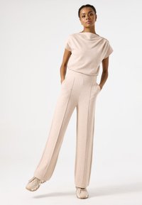 Top de punto beige con escote drapeado y mangas cortas, combinado con pantalones beige de pierna ancha y cintura alta con pliegues frontales y bolsillos laterales.