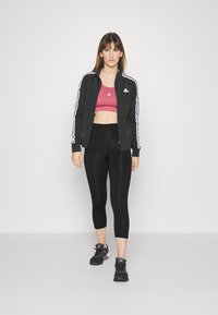 Black zip-up jacket with white stripes, pink sports bra, black leggings, and black athletic shoes. Smooth fabric with fitted design.