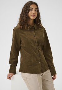 Žena s vlnitými hnedými vlasmi, oblečená v zapínanej olivovo zelenej kordovej košeli a béžových nohaviciach, opierajúca sa o biely povrch pred jednoduchým pozadím.