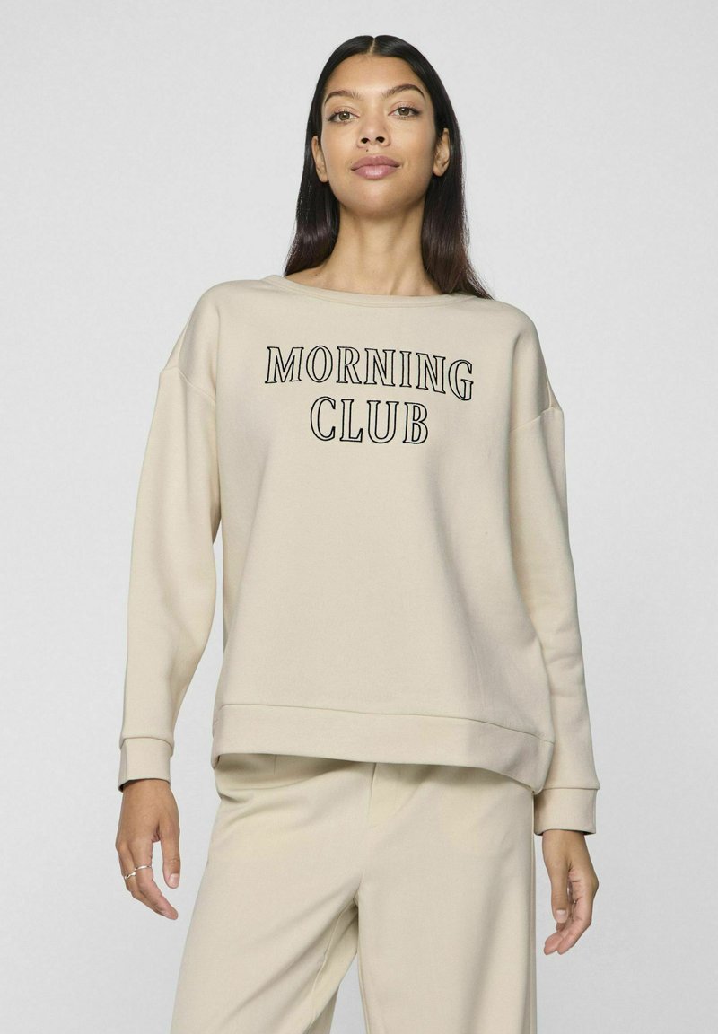 Sudadera beig con cuello redondo, mangas largas y la inscripción "MORNING CLUB" en negro. Textura suave, corte relajado, combinada con pantalones a juego.