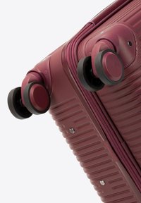 Valigia rigida color bordeaux con texture lineare, quattro ruote nere multidirezionali, doppia cerniera e dettagli in metallo.