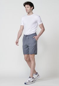 Uomo che indossa una maglietta bianca, pantaloni corti grigi con cordino e sneakers bianche, in piedi con una mano in tasca su uno sfondo semplice.