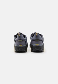 Zapatillas atléticas de color gris oscuro con suelas negras contrastantes, material texturizado y acentos amarillos. Presentan un diseño robusto para uso al aire libre.