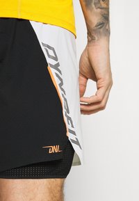 Shorts de sport noir et blanc avec des accents orange, texture perforée et détail de logo sur le côté. Un haut jaune est partiellement visible.