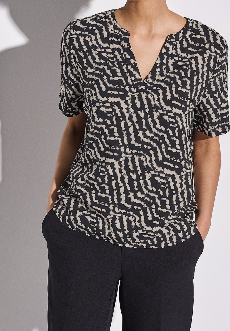 Kortärmad svart och beige mönstrad topp med V-ringning och avslappnad passform, i kombination med svarta byxor. Slät, lättvikts tyg.