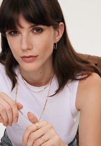 Złota naszyjnik z prostokątnymi ogniwami, nałożony na krótszy delikatny łańcuszek. Modelka nosi pasujące złote kolczyki w kształcie kół oraz pierścionki.