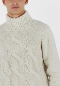 Crème-kleurige gebreide trui met een hoge ribbelkraag, kabelpatronen en textuurdetails op het lichaam.