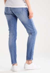 Osoba v světle modrých slim-fit džínech a bílých perforovaných teniskách, postávající před jednobarevným světlým pozadím.