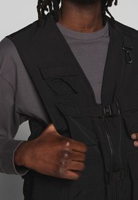 Schwarze Utility-Weste aus leichtem Stoff mit mehreren Taschen, einem Reißverschluss und einer Schnalle. Getragen über einem grauen Langarmshirt.